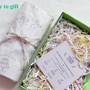 Puede incluir: Una caja de regalo verde claro llena de papel triturado y una tarjeta blanca etiquetada como Authentic Handmade Piece de Cozara Studio. Al lado, un paquete envuelto en papel blanco con ilustraciones grises de querubines y un pastel.