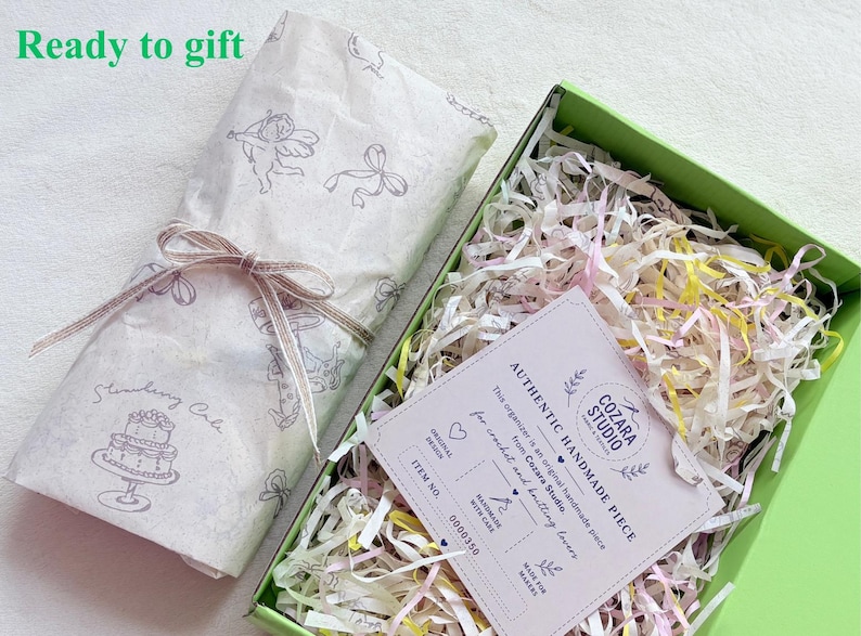 Puede incluir: Una caja de regalo verde claro llena de papel triturado y una tarjeta blanca etiquetada como Authentic Handmade Piece de Cozara Studio. Al lado, un paquete envuelto en papel blanco con ilustraciones grises de querubines y un pastel.