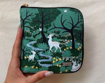Puede incluir: Un estuche cuadrado con cremallera que presenta una escena de bosque verde con ciervos blancos, un arroyo azul serpenteante, árboles y pequeñas flores. Una pequeña etiqueta metálica rectangular con el texto "Cozm" está fijada en la parte inferior derecha.