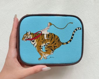 Puede incluir: Un estuche rectangular con cremallera y cubierta de tela azul claro que presenta una ilustración impresa de una persona con una trenza larga montada sobre un tigre en carrera. El tigre es marrón con rayas negras y un arnés de color rojo.