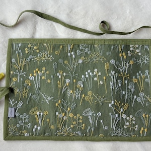 Puede incluir: Un estuche enrollable de tela verde oliva con un patr&oacute;n bordado de flores silvestres blancas y amarillas. El estuche incluye largas cintas de tela verde oliva con puntas amarillas y una peque&ntilde;a etiqueta rectangular gris con la inscripci&oacute;n "Ceotere".