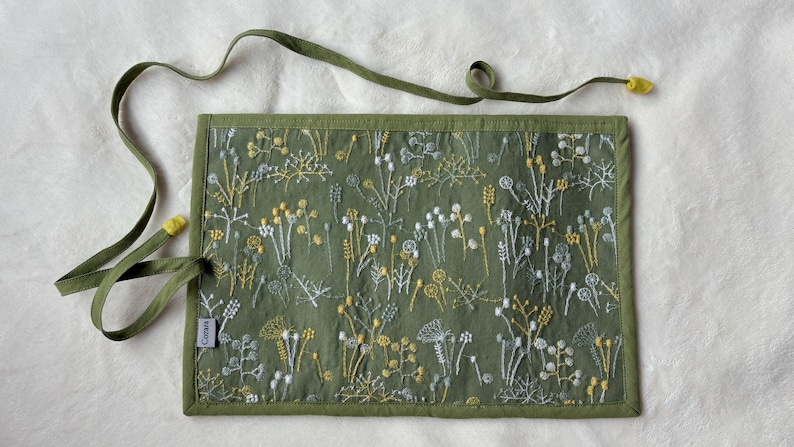 Puede incluir: Un estuche enrollable de tela verde oliva con un patr&oacute;n bordado de flores silvestres blancas y amarillas. El estuche incluye largas cintas de tela verde oliva con puntas amarillas y una peque&ntilde;a etiqueta rectangular gris con la inscripci&oacute;n "Ceotere".