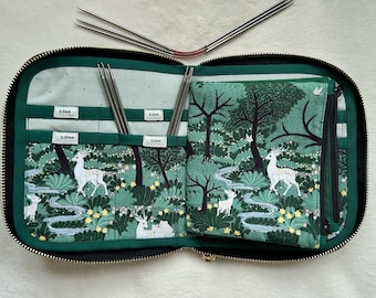 Peut inclure: Un étui en tissu zippé avec un imprimé forêt vert représentant des cerfs blancs, des arbres et des fleurs. L'intérieur comporte plusieurs poches pour aiguilles à tricoter, étiquetées 2.0mm, 2.25mm, 3.0mm et 3.5mm. Des aiguilles sont rangées.