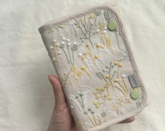 Peut inclure: Un étui rectangulaire en tissu beige orné de motifs botaniques brodés en jaune, blanc et vert sauge. L'étui comprend deux boutons-pression vert clair pour la fermeture et une petite étiquette en tissu gris avec le texte Cozara sur le côté.