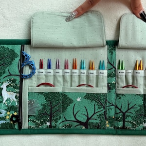 May include: A fabric knitting needle organizer case featuring a green forest print with deer and trees. The interior contains multiple slots holding various colored metal knitting needles, each labeled with its specific metric size in millimeters.