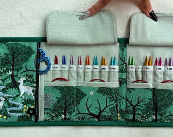 Peut inclure: Un étui de rangement en tissu pour aiguilles à tricoter avec un imprimé forêt vert représentant des cerfs et des arbres. L'intérieur comporte plusieurs fentes contenant des aiguilles en métal colorées, chacune marquée de sa taille en millimètres.