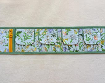 Peut inclure: Un organisateur en tissu long et rectangulaire avec un fond bleu clair orné d'un imprimé floral et d'oiseaux colorés. Il comprend une poche zippée jaune verticale sur la gauche, cinq petits compartiments à rabat et une bordure verte.
