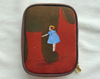 Peut inclure: Une pochette rectangulaire à fermeture éclair avec une bordure marron foncé. La face avant présente un fond rouge avec la grande silhouette marron foncé d'une créature et une petite illustration d'une personne blonde en robe bleue.