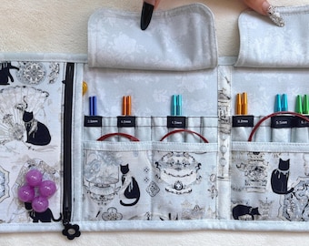 Peut inclure: Un étui de rangement en tissu pour aiguilles à tricoter avec un motif de chats noirs et blancs. L'intérieur comporte plusieurs fentes contenant des aiguilles métalliques colorées avec des tailles indiquées, une poche zippée et quatre marqueurs de mailles violets.