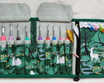 Peut inclure: Un étui en tissu pour crochets avec un motif de forêt verte, des cerfs blancs, des fleurs jaunes et un ruisseau. L'intérieur comporte plusieurs fentes contenant divers crochets en métal à poignées colorées, une poche zippée et des marqueurs de mailles.
