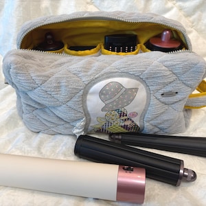 May include: A quilted light gray fabric storage bag with a yellow interior lining and a central patch featuring a person in a hat. The bag contains several small containers, and a hair styling tool with a cream handle and black attachments lies in front.