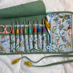 May include: A green fabric roll-up organizer featuring a light blue floral and bird print interior. It contains several colorful crochet hooks, a pair of light pink scissors, a yellow seam ripper, a white measuring tape, and four plastic stitch markers.