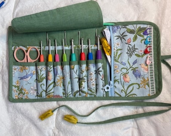 Peut inclure: Un organisateur en tissu vert à enrouler avec un intérieur à motif floral et oiseaux bleu clair. Il contient plusieurs crochets colorés, une paire de ciseaux rose clair, un découd-vite jaune, un ruban à mesurer blanc et quatre marqueurs de mailles.