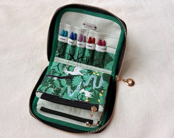 Peut inclure: Un étui en tissu zippé noir et vert, orné d'un imprimé forêt avec des oiseaux blancs. L'intérieur contient cinq aiguilles à tricoter colorées de tailles 3.0mm, 4.0mm, 4.5mm, 5.0mm et 6.0mm, rangées dans des compartiments avec une petite étiquette.