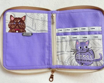 Peut inclure: Un étui zippé en tissu lavande avec une illustration de chat brun à gauche et une illustration de chat violet à droite. L'intérieur comprend des emplacements étiquetés pour des aiguilles à tricoter de 3,5 mm, 3,75 mm, 4,0 mm, 4,5 mm, 5,0 mm, 5,5 mm, 6,0 mm, 6,5 mm et 7,0 mm.