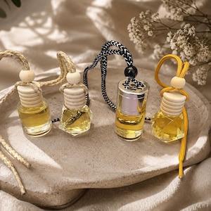 May include: Four small glass car air freshener bottles filled with golden liquid. Three bottles have faceted bases and light wooden caps with hanging cords. One bottle has a cylindrical silver cap and a black and white patterned hanging cord.