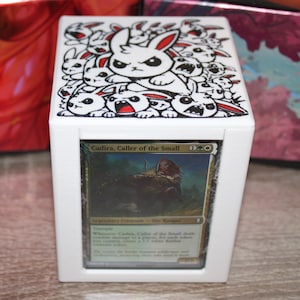 May include: White card storage box featuring a cartoon rabbit design with red accents. The box holds a card with the text "Cadira, Caller of the Small". The box is on a wooden surface.