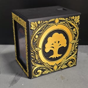 May include: A black rectangular storage box featuring a prominent gold-colored tree emblem centered within a circular frame. The box is decorated with intricate gold-colored filigree and leaf patterns along the edges and corners of the front panel.