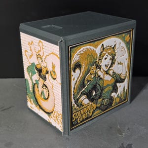 May include: A gray 3D-printed storage box featuring a rectangular design with two illustrated panels. The front panel displays a comic-style character with a squirrel tail and the text "The Unbeatable SQUIRREL GIRL". The side panel shows a character.