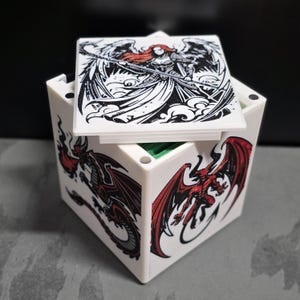 May include: A white square storage box featuring black and red graphic illustrations on each side. The lid displays a winged figure holding a staff, while the sides show a red horned demon with wings and a black dragon-like creature with red accents.