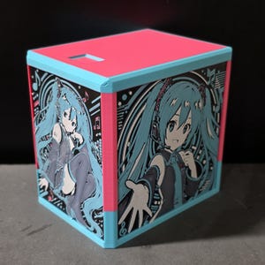 May include: A cubic storage box featuring a bright pink top and light blue edges. The side panels display a black and light blue graphic illustration of an anime character with long teal hair, headphones, and musical notes, set against a circular background.