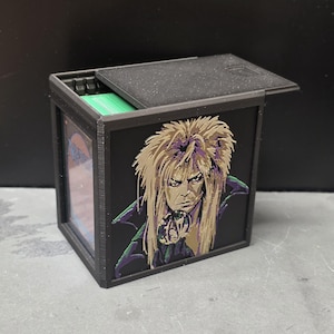 May include: A black rectangular deck box with a sliding lid, featuring a detailed graphic of a person with long blonde hair holding a crystal ball on the front panel. The box contains a stack of green-edged cards visible through the open top.