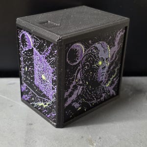 May include: A dark gray, cube-shaped storage box featuring a textured, 3D-printed design on its side panels. The artwork displays a purple and yellow graphic of a robotic figure and a geometric structure against a dark, starry space background.