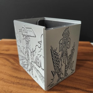 May include: A silver-colored, square-shaped container with etched illustrations of a character from a video game. The artwork depicts a figure with spiky hair and a sword, set against a backdrop of buildings and a road.