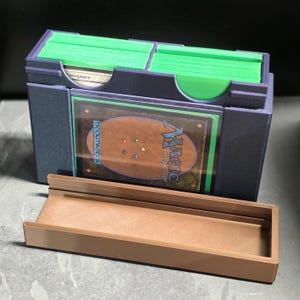 May include: A dark purple plastic trading card storage box with two compartments holding stacks of cards in bright green sleeves. A Magic: The Gathering card is displayed in a clear front slot. A separate, empty brown plastic tray sits in front.