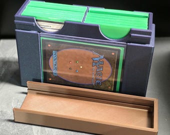 May include: A dark purple plastic trading card storage box with two compartments holding stacks of cards in bright green sleeves. A Magic: The Gathering card is displayed in a clear front slot. A separate, empty brown plastic tray sits in front.