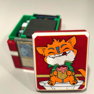May include: A red and white card case with a cartoon fox illustration holding a mug. The case is open, revealing a green and red card storage box filled with cards. The case has a gold border.