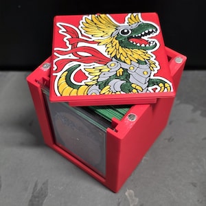 May include: A red square deck box for trading cards with a magnetic lid. The lid features a colorful illustration of a green, yellow, and silver armored dinosaur-like creature with a red cape. The box contains a stack of cards with green edges.