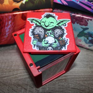 May include: A red square box with a magnetic lid featuring a green goblin illustration. The goblin has pointed ears, wears a fur-trimmed collar, and holds a white orb. The box interior contains dark green stacked cards visible beneath the lid.