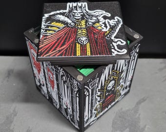 May include: A black square storage box with a removable lid featuring a graphic of a skeletal king in a red cape and crown. The sides of the box display detailed black and white illustrations of gothic architecture, thrones, and banners with skull motifs.