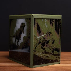 May include: A green cube-shaped decorative box featuring dinosaur illustrations. The box has a dark green frame and depicts a Tyrannosaurus Rex and other dinosaurs in a forest setting. The box is displayed on a wooden surface.
