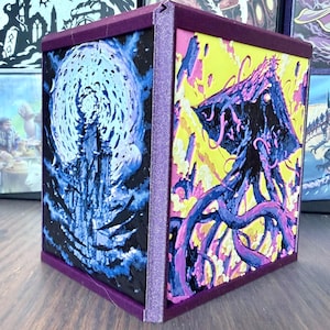 May include: A square decorative box with a purple frame. One side features a blue and black illustration of a tall structure under a large moon. The adjacent side displays a purple and black tentacled creature against a bright yellow background.