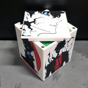 May include: A white square storage box featuring black and white anime-style character illustrations with red and blue accents on the sides and lid. The box has a removable lid and a green interior, with small circular magnets visible on the top corners.