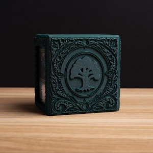 May include: A dark teal, cube-shaped box with ornate, embossed detailing. The front features a circular emblem with a tree design. The box is set on a light wood surface against a black backdrop.