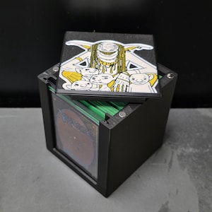 May include: A black cubic card storage box with a magnetic lid featuring a graphic illustration of a large goblin-like creature with long hair and three smaller goblin figures. A Magic: The Gathering trading card is visible inside the box.