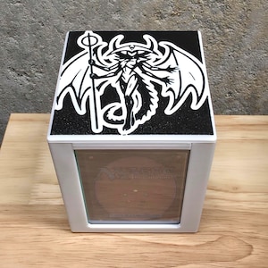 May include: White card storage box with a black top featuring a white graphic of a fantasy figure with wings and a staff. The box has a clear front panel and is sitting on a wooden surface.