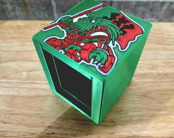 May include: A green, cube-shaped box with a black interior. The top of the box features a colorful illustration of a creature holding a spear and a red flag. The box is sitting on a wooden surface.