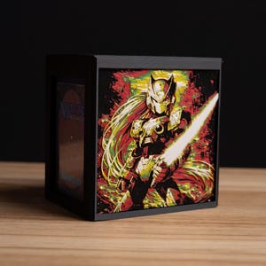 May include: Black wooden card storage box featuring a colorful illustration of a character with a glowing sword. The artwork has red, yellow, and green accents. The box is designed to hold trading cards.