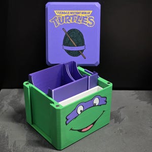 May include: A green and purple storage box featuring a Teenage Mutant Ninja Turtles graphic on the lid and a Donatello face design on the front. The box includes internal purple dividers and a white tray, with visible magnetic closures on the corners.