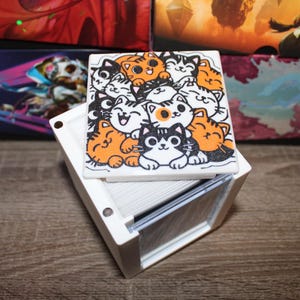 May include: A white card storage box with a lid featuring a cartoon illustration of several cats in various poses and colors. The cats are orange, black, and white. The box is sitting on a wooden surface.