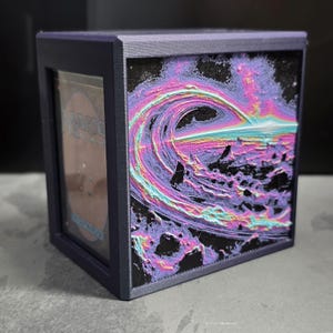May include: A dark purple card display box with a colorful, abstract wave design on the front. The wave features shades of pink, purple, and blue against a black background. The box has a clear panel to view the card.