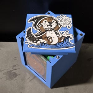 May include: A light blue square storage box with a removable lid featuring a brown and white cartoon otter wearing a hooded cape, splashing in water. The box has a clear window on the side revealing a stack of cards inside with metallic fasteners.