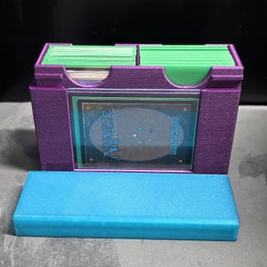 May include: A purple plastic card storage box with two compartments holding stacks of green-sleeved trading cards. The front features a clear window displaying a Magic The Gathering card back. A separate, solid blue rectangular lid sits in front.