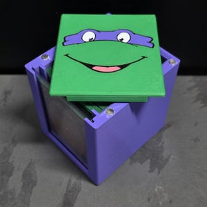 May include: A purple square storage box with a green lid featuring a graphic of a cartoon turtle face wearing a purple mask. The box has a clear side panel and metallic silver fasteners at the corners, containing a stack of green-edged cards.