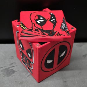 May include: A red square storage box with a removable lid featuring a black and red graphic of the character Deadpool. The side of the box displays a full-body cartoon illustration of the character and a large circular Deadpool logo in black.