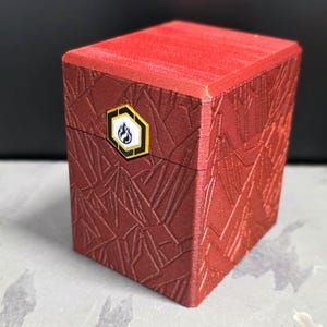 May include: A red, textured box with a geometric pattern and a hinged lid. The box features a gold and black emblem with a flame design. The box is likely for storing small items.